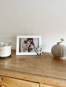 Witte fotolijst met 3D-panda van Deknudt op een houten dressoir in een huiselijke setting. In de lijst zit een foto van een moeder met haar twee kinderen. De lijst staat tussen een geurkaars en een vaasje, wat zorgt voor een warme sfeer in de kinderkamer of woonkamer.