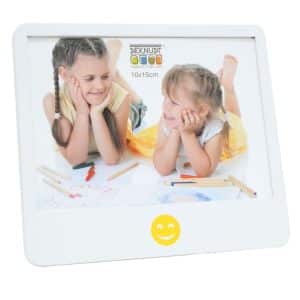 Vooraanzicht van een speelse witte fotolijst van Deknudt met een vrolijke gele smiley centraal onder het fotovlak. In de lijst zit een kleurrijke kinderfoto van twee meisjes die liggend tekenen en elkaar lachend aankijken.