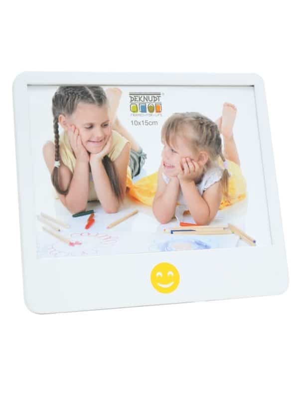 Vooraanzicht van een speelse witte fotolijst van Deknudt met een vrolijke gele smiley centraal onder het fotovlak. In de lijst zit een kleurrijke kinderfoto van twee meisjes die liggend tekenen en elkaar lachend aankijken.