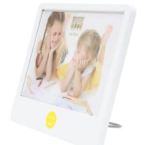 Schuin zijaanzicht van een witte Deknudt fotolijst van 10x15 cm met een gele smiley onderaan. In de lijst zit een foto van twee jonge meisjes die lachend naar elkaar kijken terwijl ze tekenen. De lijst staat stabiel op een metalen standaard.